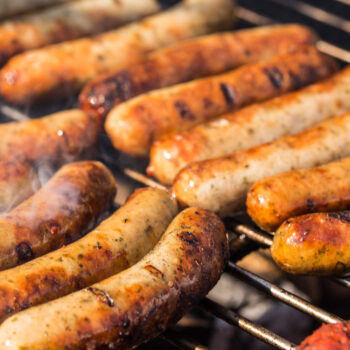 Fresh Lincolnshire sausages sizzling and browning on a barbecue grill, made with Newly Weds Colourex Lincolnshire Sausage Seasoning from Parkers Food Machinery Plus.