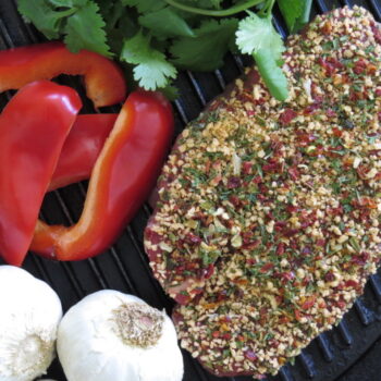 Leonards Gluten Free Pepper Steak Coater 2kg Tub seasoning applied to steak with garlic, red bell peppers, and fresh herbs on a grill pan