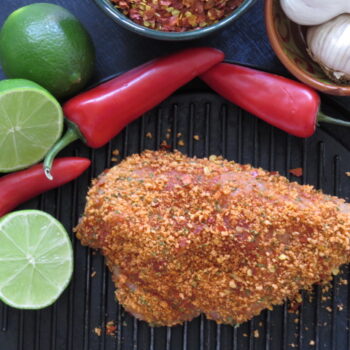 Seasoned meat coated with Leonards Gluten Free Sweet Chilli and Lime Coater, surrounded by fresh limes, chili peppers, garlic, and crushed red pepper