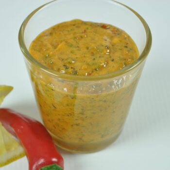 A glass of Raps Magic Marinade Lemon and Chili sauce, with a lemon wedge and red chili on a white background, a product from Parkers Food Machinery Plus.