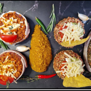 Gourmet meat portions topped with cheese, tomato, and herbs, some wrapped in bacon, arranged with garlic, chili, and herbs on dark marble showcasing Leonards Firey Crumb Coater seasoning blend, available from Parkers Food Machinery Plus