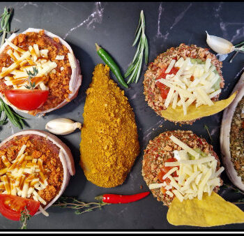 Gourmet meat portions topped with cheese, tomato, and herbs, some wrapped in bacon, arranged with garlic, chili, and herbs on dark marble showcasing Leonards Firey Crumb Coater seasoning blend, available from Parkers Food Machinery Plus