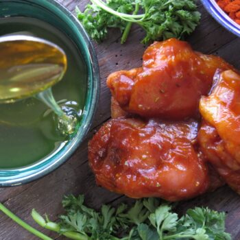 Sticky maple glaze being poured into a bowl beside barbecue-coated chicken, fresh parsley, and paprika