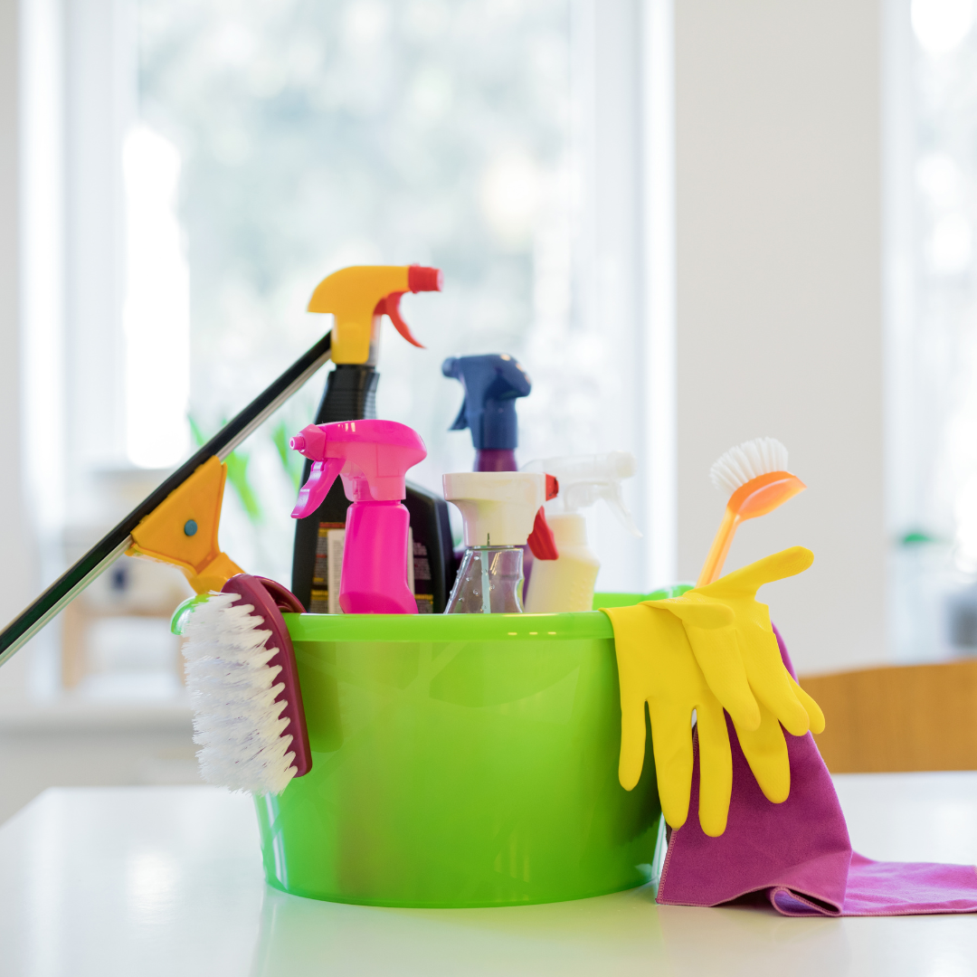 Green bucket filled with assorted cleaning supplies including spray bottles, brushes, gloves, and cloths in a bright indoor setting