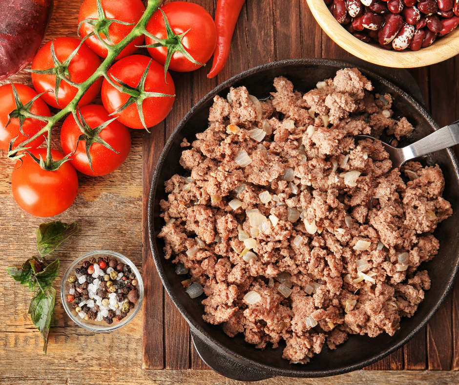 Fresh tomatoes, red beans, chili pepper, peppercorns, and cooked ground meat with onions in a skillet arranged on a wooden surface for meat mincer preparation