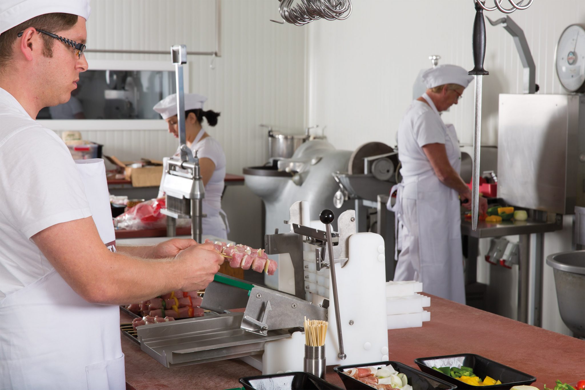 Commercial kitchen staff preparing meat and vegetable skewers using PINTRO P160 skewer machine, with food trays and processing equipment in view