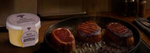A tub of Black Country Beef Dripping next to a hot frying pan where three thick-cut steaks are being seared to a perfect golden-brown crust.