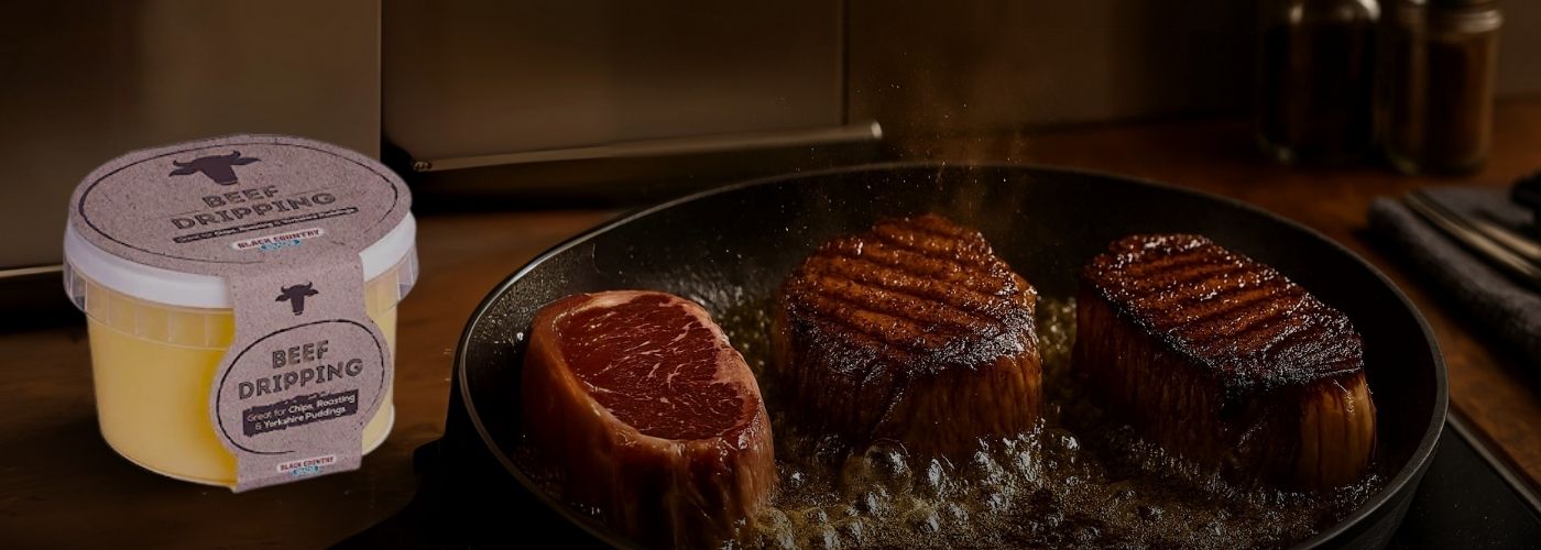 A tub of Black Country Beef Dripping next to a hot frying pan where three thick-cut steaks are being seared to a perfect golden-brown crust.