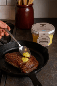 A hand spooning dollops of Black Country Beef Dripping onto a freshly seared steak in a cast-iron skillet to add flavor.