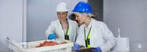 Butchers wearing protective PPE inspecting fresh minced meat in a food processing area, demonstrating safe hygiene practices.