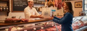 A friendly butcher in a white uniform hands a happy customer a branded paper counter bag over a meat display case in a classic butcher shop.