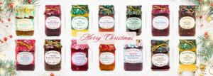 A festive banner featuring a full range of Mrs Darlington’s Christmas preserves, chutneys, curds, marmalades, and sauces. Each jar is decorated with colourful seasonal fabric covers and gold ties, arranged in a single row with a ‘Merry Christmas’ message in elegant red script across the centre. Background includes subtle Christmas greenery, baubles, and glitter for a holiday theme at PFM Plus.